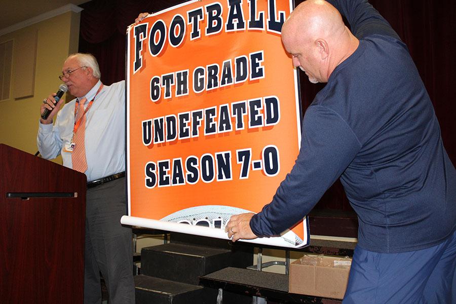 Mr. Harbeck (left) and Mr. Keller unfurl the banner awarded to the sixth-grade football team for their undefeated season.